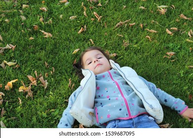 Little Girl Lying On Green Grass And Looking At The Sky. Snow Angel On The Lawn With Fallen Leaves.