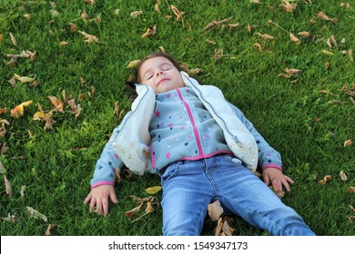 Little Girl Lying On Green Grass And Looking At The Sky. Snow Angel On The Lawn With Fallen Leaves.