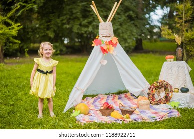Little girl lying and playing in a tent, children's house wigwam in park Autumn portrait of cute curly girl. Harvest or Thanksgiving. autumn decor, party, picnic. Child in yellow dress with apples.