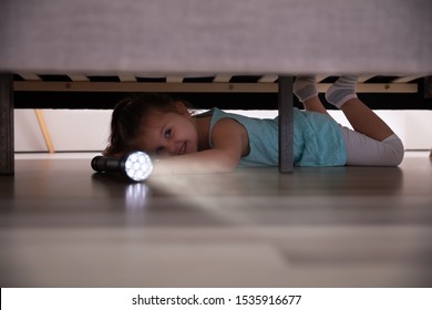 Little Girl Lying Under The Bed Searching For Something With Flash Light At Home