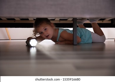 Little Girl Lying Under The Bed Searching For Something With Flash Light At Home