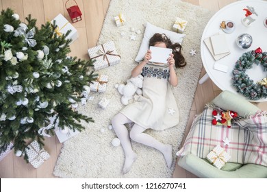 Little Girl Is Lying Under The Christmas Tree With A Tablet On The Carpet. View From Above