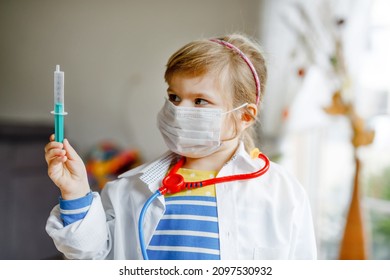 Little Girl Makes Injection. Cute Child With Medical Mask Playing Doctor, Holding Syringe With Vaccine. Coronavirus Covid Vaccination Concept. Kid Play Role Game At Home.