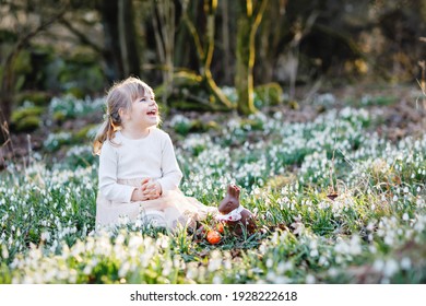 Little Girl Making Easter Egg Hunt In Spring Forest On Sunny Day, Outdoors. Cute Happy Child With Lots Of Snowdrop Flowers And Chocolate Bunny. Springtime, Christian Holiday Concept.