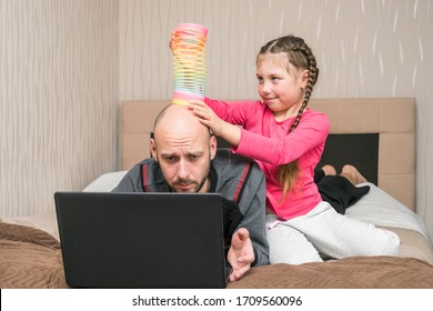 A little girl is making fun of dad.A child prevents a man from working on a laptop in his bedroom. The daughter puts the slinky toy to the father's head. Family is quarantined at home.