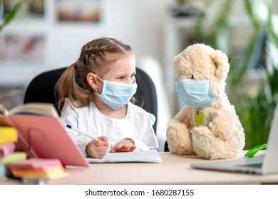 little girl in mask, with Teddy bear, doing homework, writing in notebook, using laptop, e-learning concepts, during quarantine and self- isolation. e-learning due to Covid-19 coronavirus pandemic.