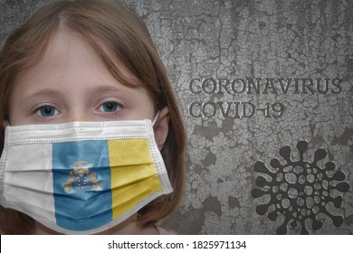 Little Girl In Medical Mask With Flag Of Canary Islands Stands Near The Old Vintage Wall With Text Coronavirus, Covid, And Virus Picture.