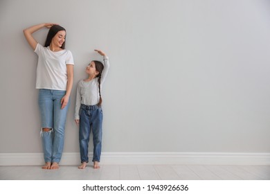 Little girl and mother measuring their height near light grey wall indoors. Space for text