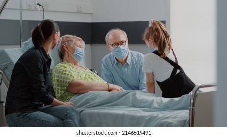 Little girl and mother visiting senior woman with face mask in hospital ward bed. Child and adult talking to ill grandma at family visit during coronavirus pandemic. Visitors with sick patient