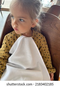 Little Girl With A Napkin Tucked Into Her Collar Sits In A Restaurant