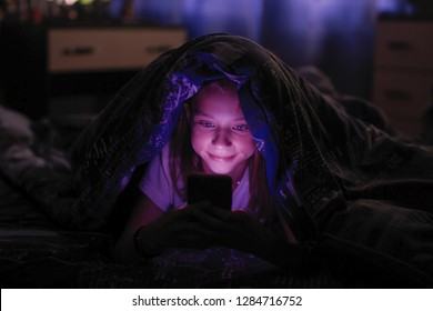 Niña pequeña por la noche mira el smartphone bajo una manta en la cama.