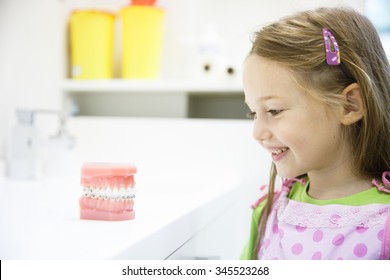 Little girl observing artificial model of human jaw with dental braces in dentists office, smiling. Pediatric dentistry, aesthetic dentistry, early education and prevention concept. 