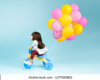 Little Girl On A Kids Tricycle With Colorful Balloons Behind On Plain Background.