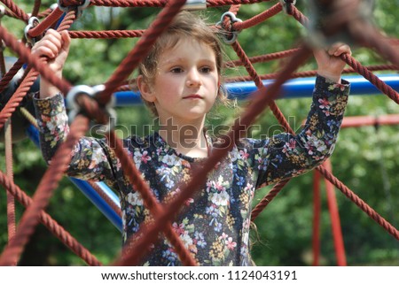 The little girl on the playground surrounded by ropes. The playground is located in the park between green trees.