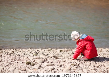 Little girl on the river collects pebbles. A child in a red jacket sat by the river.