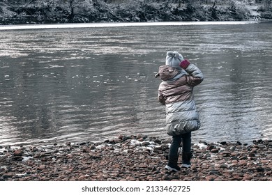 A Little Girl On A Winter Walk Near A River With A Fast Current Throws Stones At Her And Rejoices