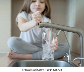Little girl opens a water tap with her hand holding a transparent glass. Kitchen faucet. Glass of clean water. Filling cup beverage. Pouring fresh drink. Hydration. Healthcare. Healthy lifestyle