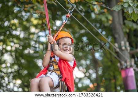 A little girl in an orange helmet and special protection goes down the zip line in a rope amusement park, at children's leisure, at a playground for toddlers, at an amusement park.
