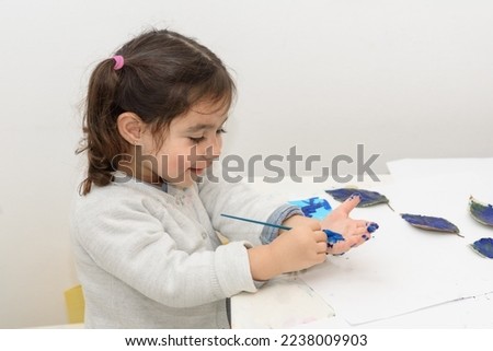 Little girl painting leaves blue color, crafts and art therapy. Child paints her own hand with blue paint and paintbrush getting messy and having fun.