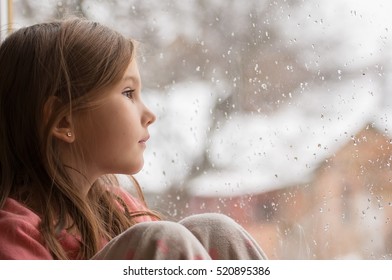 Little Girl In Pajamas Looking Out The Window At The First Snow