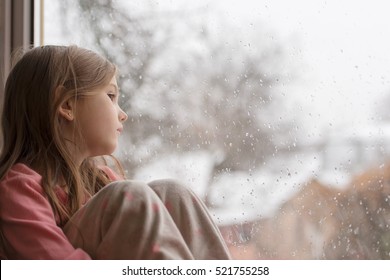 Little Girl In Pajamas Looking Out The Window At The First Snow