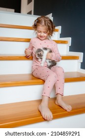 little girl in pajamas is sitting at home on the stairs playing with a kitten