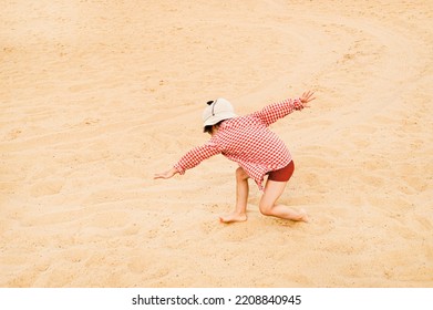 A Little Girl In A Panama And A Red Plaid Shirt Does Exercises, Stretches, Movements On A Sandy White Beach. The Child Is Dancing Breakdance. Children And Nature. Summer Vacation, Postcard. Summer