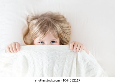 Little girl peeking out blanket on bed. Cute kid smiling and hiding under knitted cover. Palyful and mischievous eyes. Hide-and-seek. Children having fun playing active games. Happy childhood concept