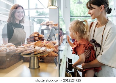 Little girl picking herself some croissants, pointing with index finger, while mother holding her in hands. Cashier smiling at them. Side view. - Powered by Shutterstock