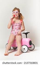 Little Girl With Pigtails, Beach Goggles, Eat A Lollipop In Pink And Fuchsia Costume With Unicorns. Next To It A Vintage Toy Car In Pink Sheet Metal. Full-length Photo On A White Background