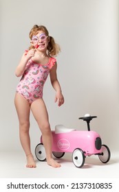 Little Girl With Pigtails, Beach Goggles, Eat A Lollipop In Pink And Fuchsia Costume With Unicorns. Next To It A Vintage Toy Car In Pink Sheet Metal. Full-length Photo On A White Background