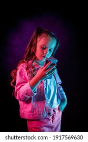 A Little Girl With Pigtails Is Looking At The Phone. Studio Photography With Neon Light, Gel Filter - Turquoise, Magenta, Purple.