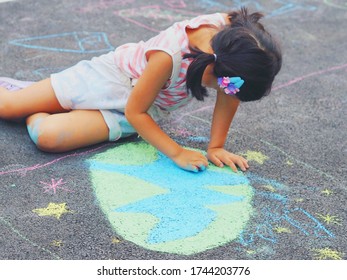 Little Girl With Pigtails Sits And Uses Colorful Chalks Draws And Paint Green Earth On The Road Floor. Earth Day Concept. Kids Outdoor Activities Concept.