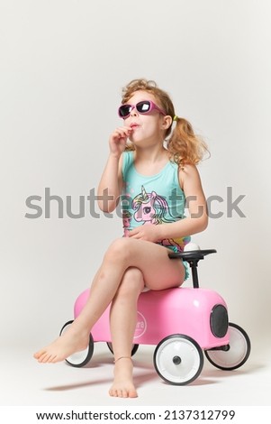 Little girl with pigtails, sunglasses, eats a lollipop in teal costume with pink unicorn. Sitting on a vintage car pink sheet metal toy. Full length photography on white background.