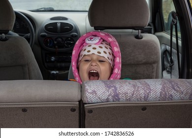 A Little Girl In Pink Clothes Screams In The Back Seat Of A Passenger Car. Portrait Of A Joyfully Screaming Girl In A Car. Travel Concept With Kids, Child Traveling By Car.