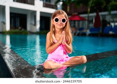 Little girl in pink dress and sunglasses sits in lotus pose by poolside. Child practices yoga, namaste gesture, enjoys tropical vacation. Wellness, family holidays concept with kid relaxing. - Powered by Shutterstock