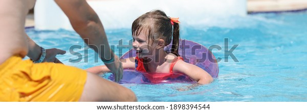 Little girl in pink lifebuoy swims to her father\
in the pool of water park. Safety rules for the behavior of child\
on water.