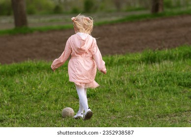 Little girl in pink outfit kicks ball in meadow. Back view. Child plays with the ball outdoors - Powered by Shutterstock