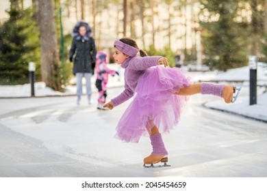 little girl in a pink sweater and a full skirt rides on a sunny winter day on an outdoor ice rink in the park
