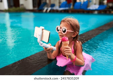 Little girl in pink swimsuit sits in pool, holds phone on selfie stick, eats ice cream, records video. Child influencer enjoys summer, vlogs lifestyle, shares content. Kid in sunglasses, water fun. - Powered by Shutterstock