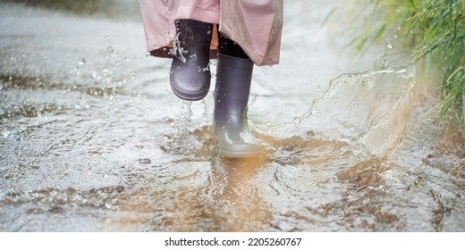 Little Girl In Pink Waterproof Raincoat, Purple Rubber Boots Funny Jumps Through Puddles On Street Road In Rainy Day Weather. Spring, Autumn. Children's Fun After Rain. Outdoors Recreation, Activity.