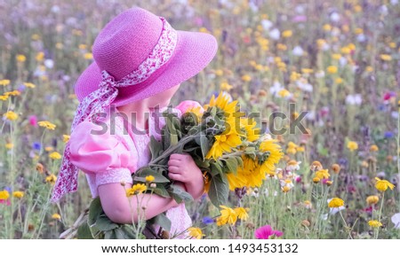 A little girl in a pink, wide-brimmed hat and pink dress holds a bouquet of sunflowers in her hands. Girl with a bouquet of flowers on a background of a meadow with meadow flowers.