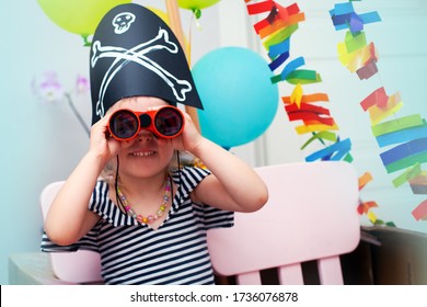 A little girl in a pirate costume looks through binoculars directly at the camera. Birthday in the style of pirates. A child in a pirate hat smiles and looks at the camera, close-up.