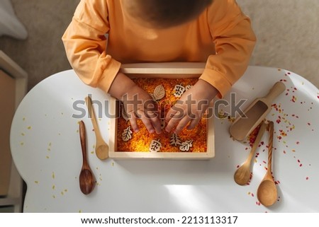A little girl playing with colored rice in autumn sensory bin. Montessori material. Scooping and pouring activity for kids. Games for learning and development of the child.