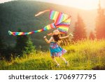 Little girl playing with colored striped rainbow kite at sunset summer day against background of beautiful mountain landscape with peaks, fir trees. Baby wearing hat and dress. Happy summer vacation