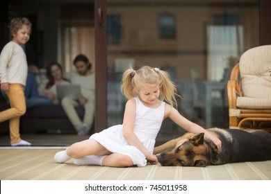 Little Girl Playing With Dog Outside, Stroking German Shepherd Lying On Porch, Parents With Small Boy Spending Time Inside Home At Background, Leisure Of Cozy Warm Happy Family In Country House