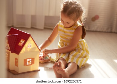 Little Girl Playing With Doll House While Siiting On Floor In Bedroom. Role Game For Young Children. Female Child Putting To Bed Her Toy Doll, Charming Little Lady Wears Yellow Dress. Children Concept