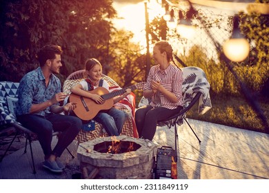 Little girl playing guitar by the fireplace, mom and dad clapping and singing along. Beautiful scene of family enjoying sunny summer day outdoors in the yard. Lifestyle, togetherness, family concept. - Powered by Shutterstock
