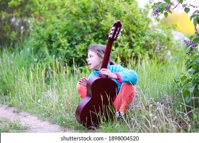 Little Girl Playing Guitar And Singing Outdoors On Green Meadow At Spring. Happy Child With Musical Instrument On Rural Yard.