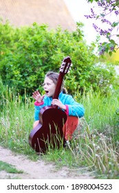 Little Girl Playing Guitar And Singing Outdoors On Green Meadow At Spring. Happy Child With Musical Instrument On Rural Yard.